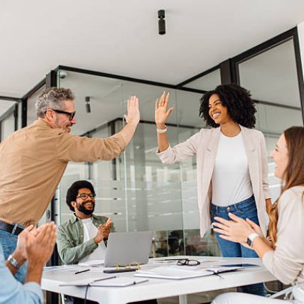 A diverse team of professionals celebrates a workplace achievement with a high-five in a modern office. The moment captures motivation, cooperation, and a positive business culture focused on teamwork
